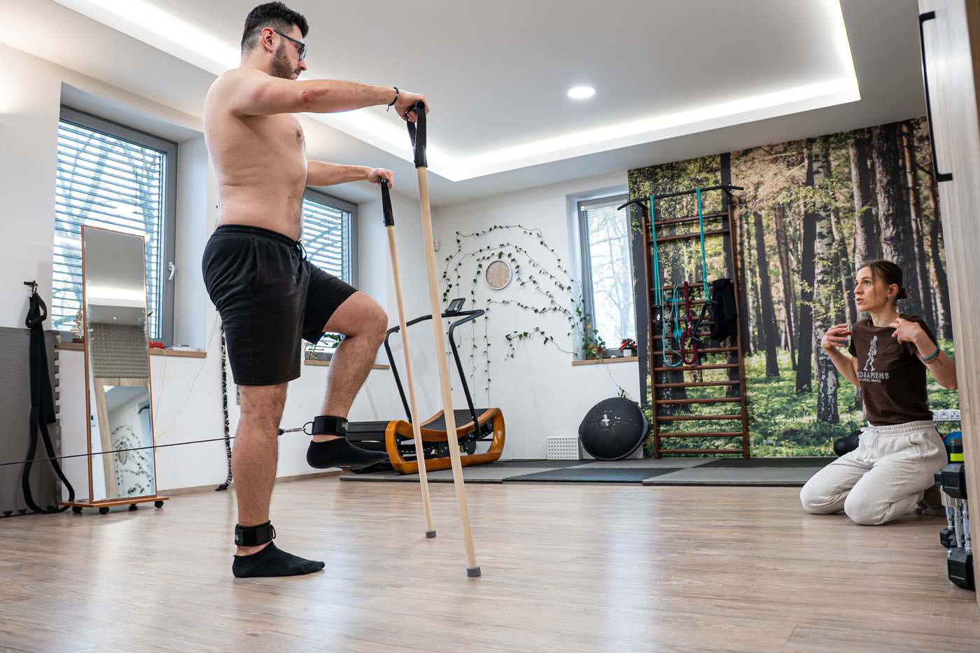 Klient ve stoji na jedné noze s holí, trenérka sleduje z podložky